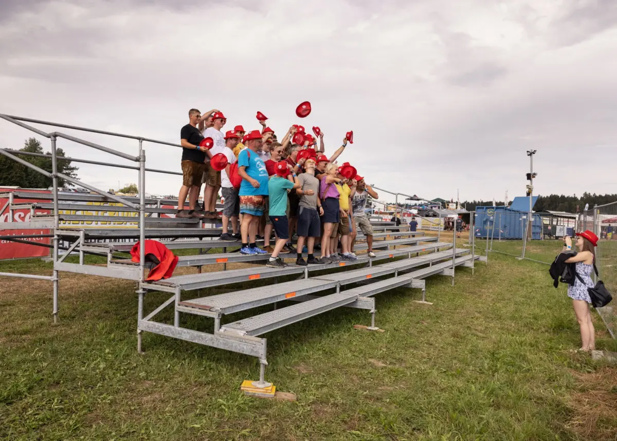 Group of fans on metal bleachers at an outdoor event, tossing red hats into the air amid banners and tents.