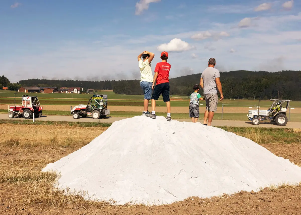 Group of four standing on a large white mound in a rural field, watching; farm buildings and tractors in the distance under a blue sky.