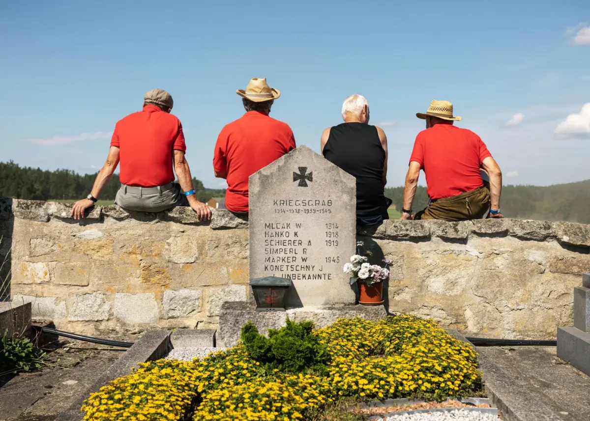 Four elderly men in red shirts and hats sit on a stone wall beside a war memorial stone with names, surrounded by yellow flowers.