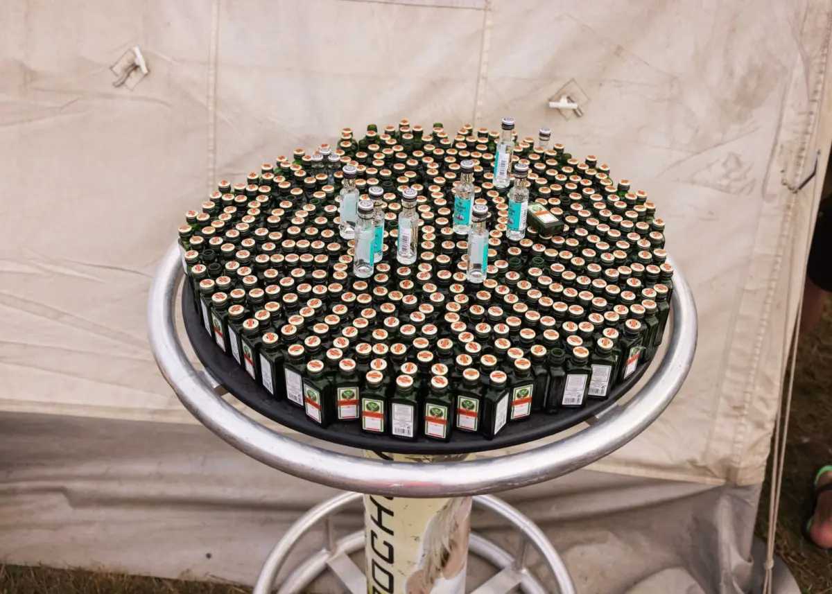 Dense pyramid of green glass bottles on a round metal table at an outdoor event under a beige tent.