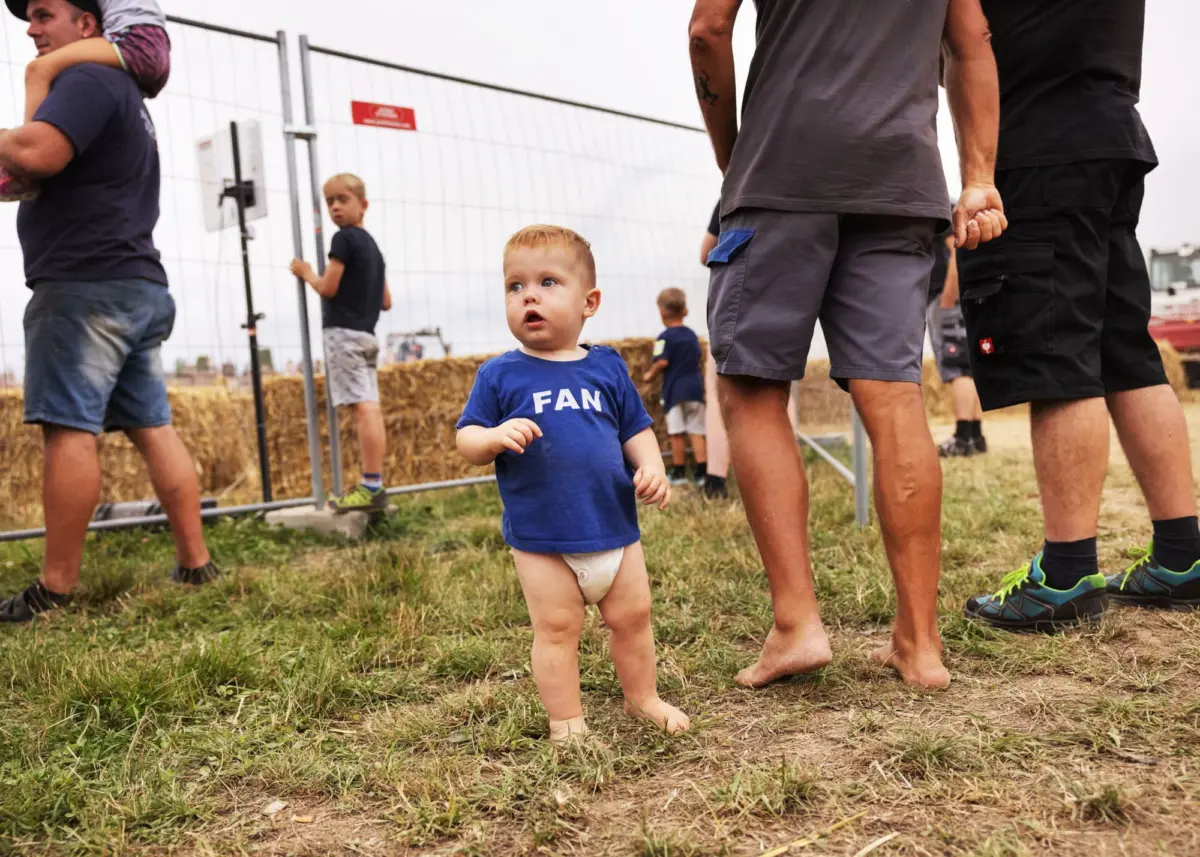 Baby in blue shirt with 'FAN' stands barefoot in a diaper on grass near a fence and hay bales at an outdoor event.