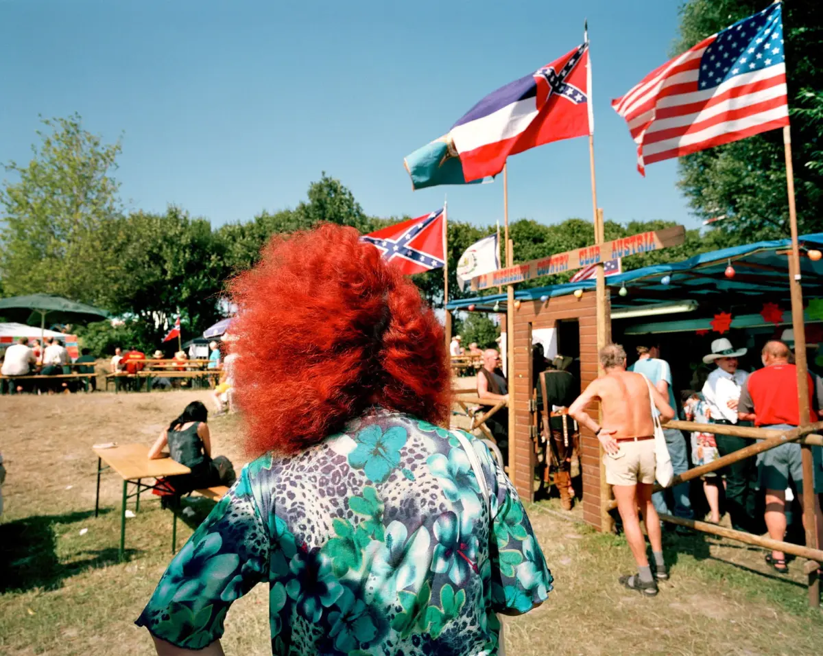 Red-haired person in a blue floral shirt seen from behind at an outdoor festival with flags and booths.