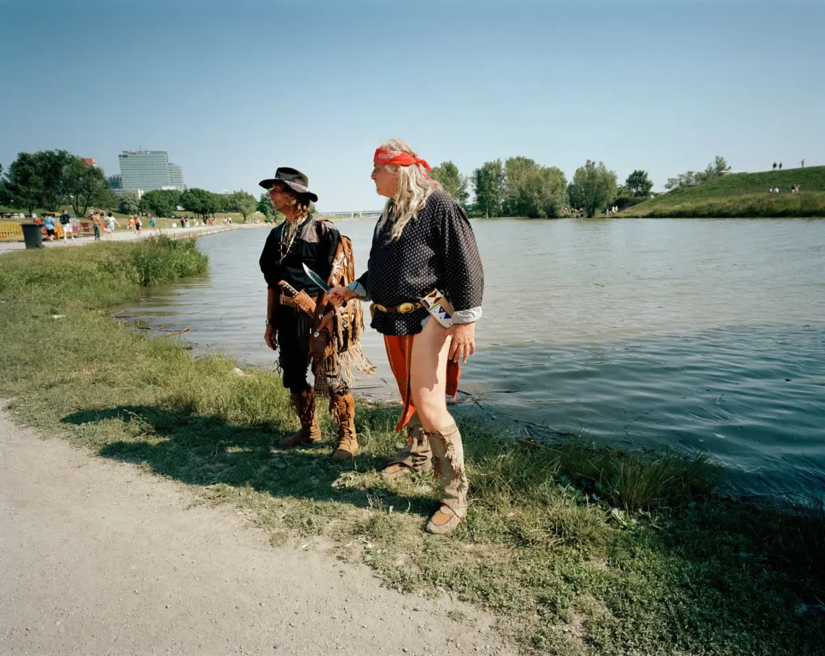 Two people in cowboy outfits stand by the riverbank, looking to the left, with a grassy park and distant buildings in the background.