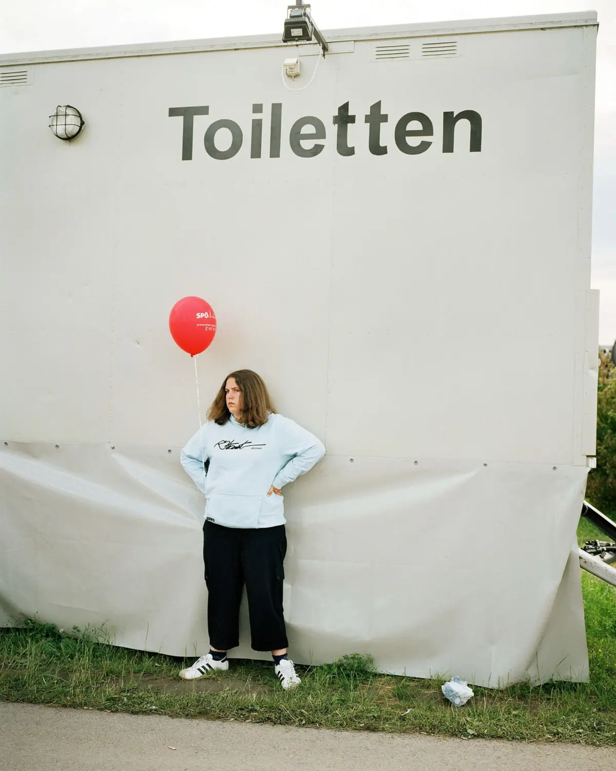 Person in light blue sweatshirt stands beside a large white trailer labeled Toiletten, holding a red balloon outdoors.