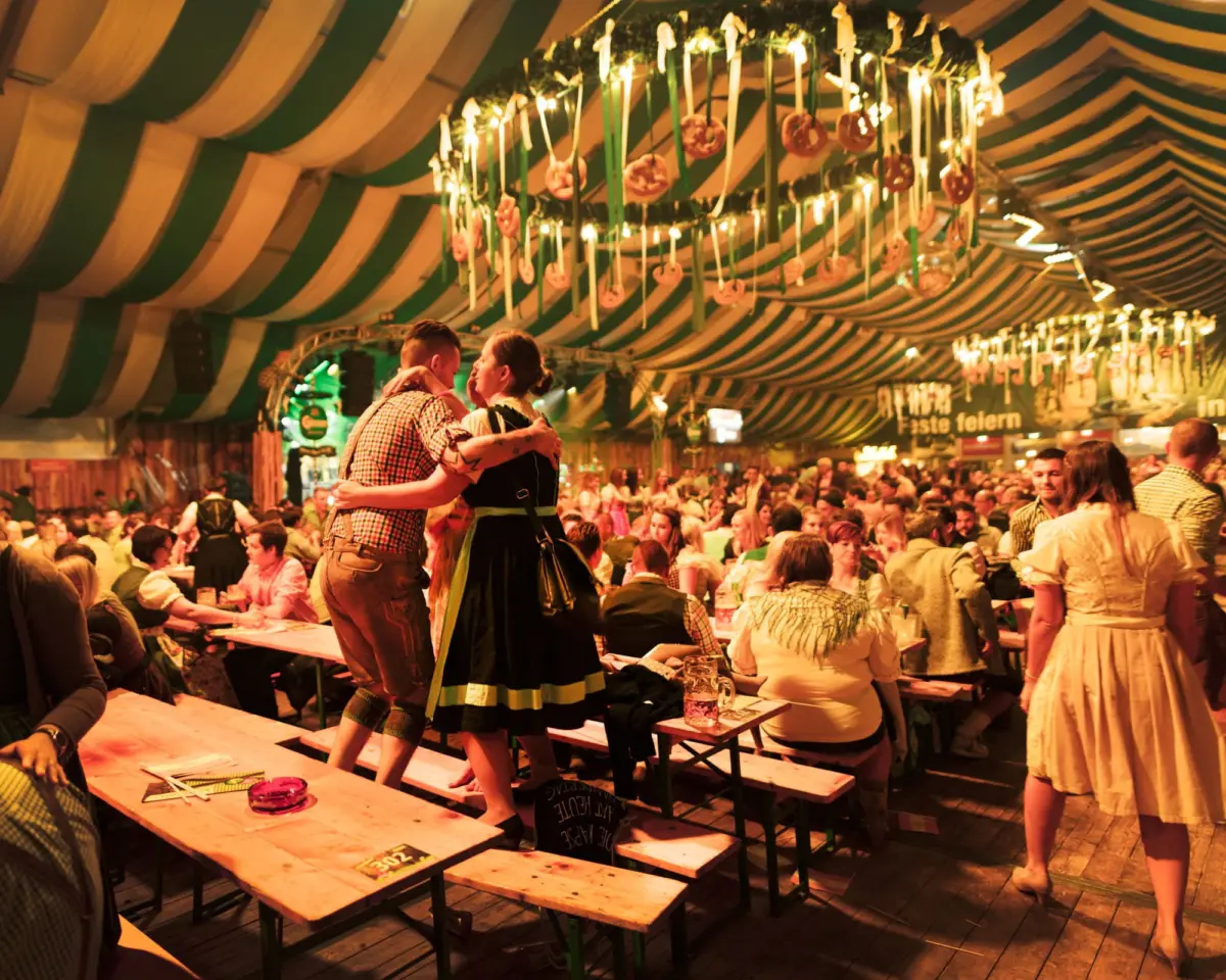 Couple in lederhosen and dirndl dancing on wooden benches inside an Oktoberfest-style beer tent.