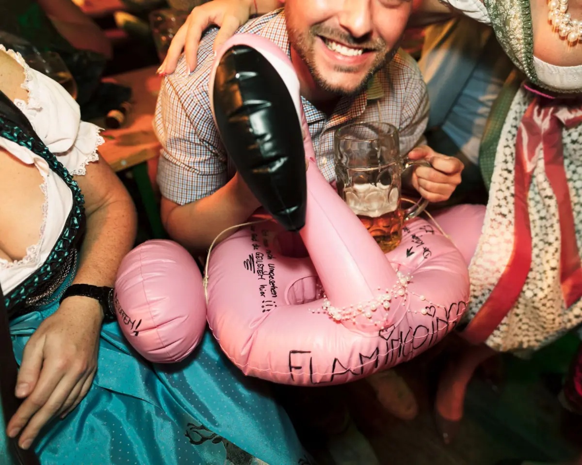 Smiling person in a checkered shirt with a pink inflatable flamingo, holding a beer mug at a festive gathering with others in traditional dress.