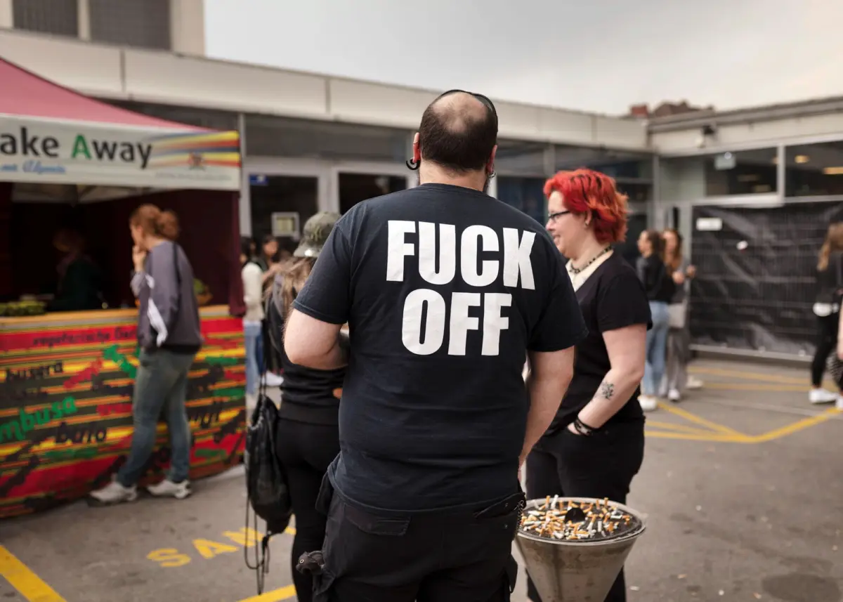 Person facing away wearing a black T-shirt with bold FUCK OFF text at an outdoor market beside a colorful vendor stall.
