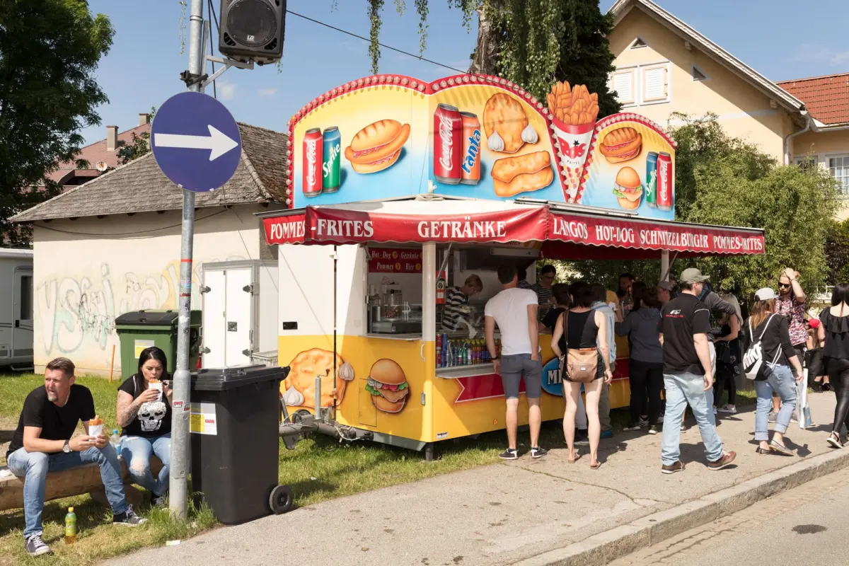 Outdoor food stall with red canopy serving pommes frites and beverages; customers queue and eat.