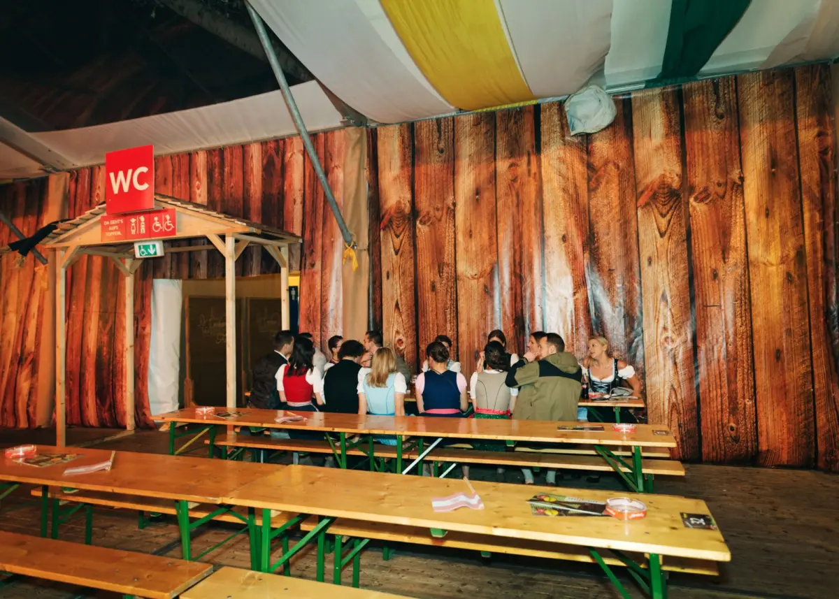 Group of people in dirndl attire socializing at long wooden tables inside a rustic hall with wood panel walls and a red WC sign.