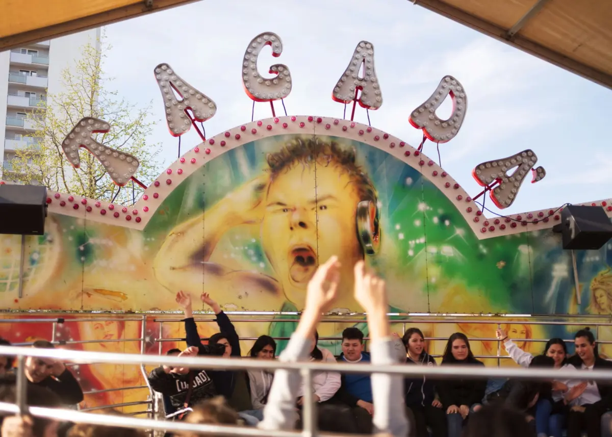 Group of teenagers on a carnival ride, hands raised, in front of a colorful mural with oversized lit letters.