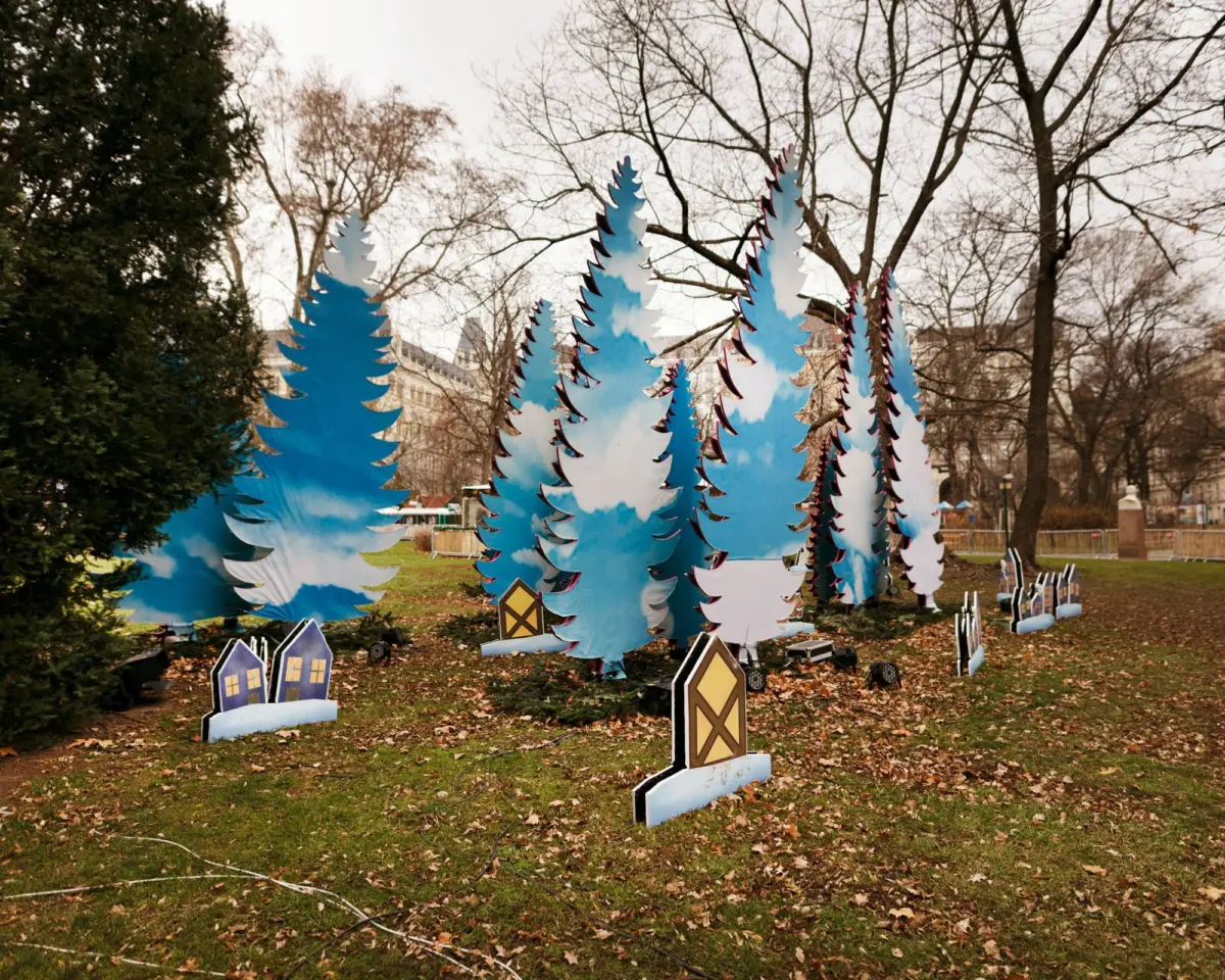 Blue stylized evergreen cutouts form an art installation in a park amid bare trees and fallen leaves.