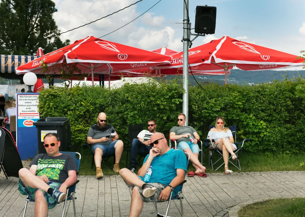 Adults lounging in folding chairs under red umbrellas at an outdoor cafe beside a hedge.