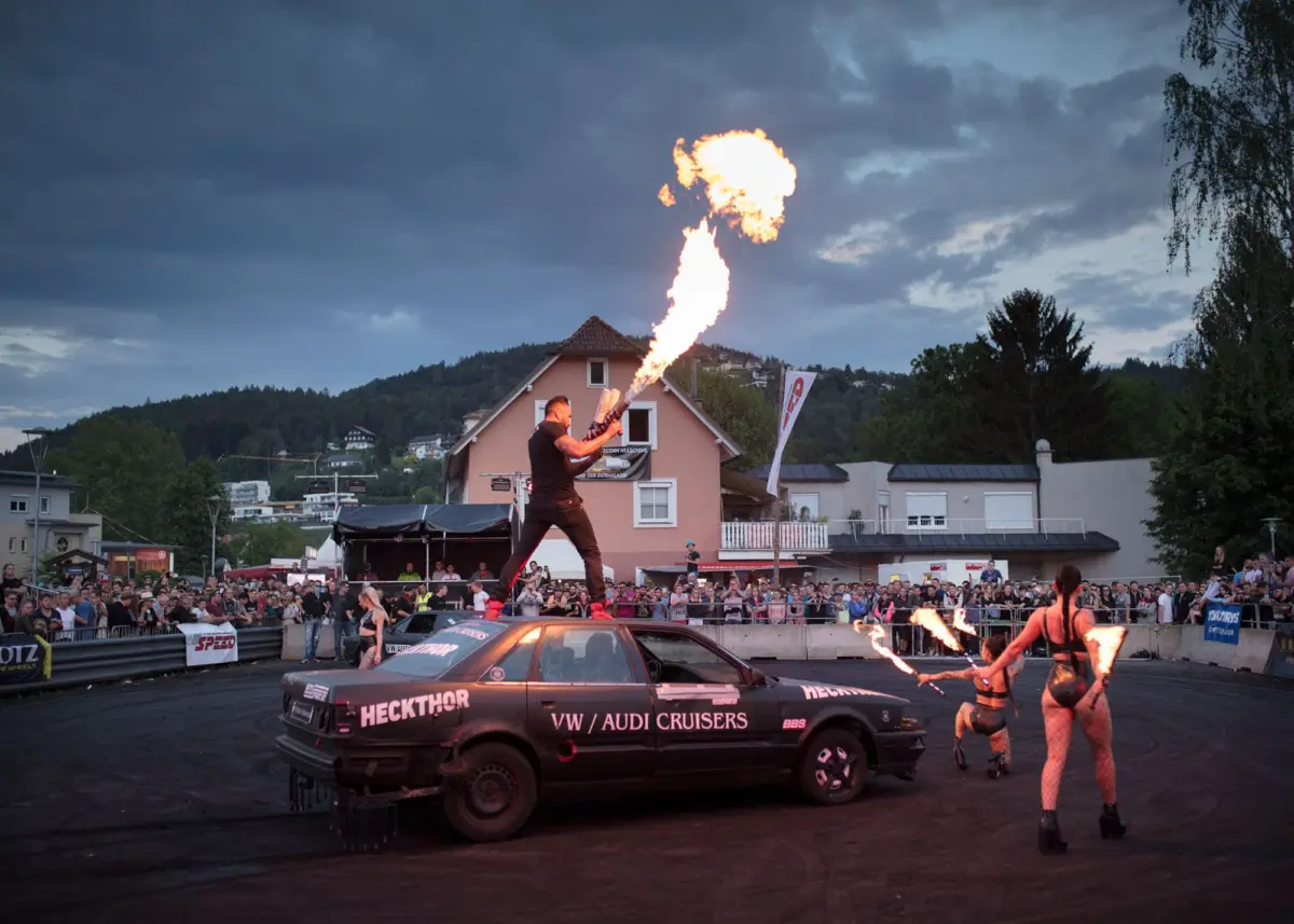 Fire performer standing on a car roof, producing a tall flame with burning torches, as a crowd watches an outdoor street festival.