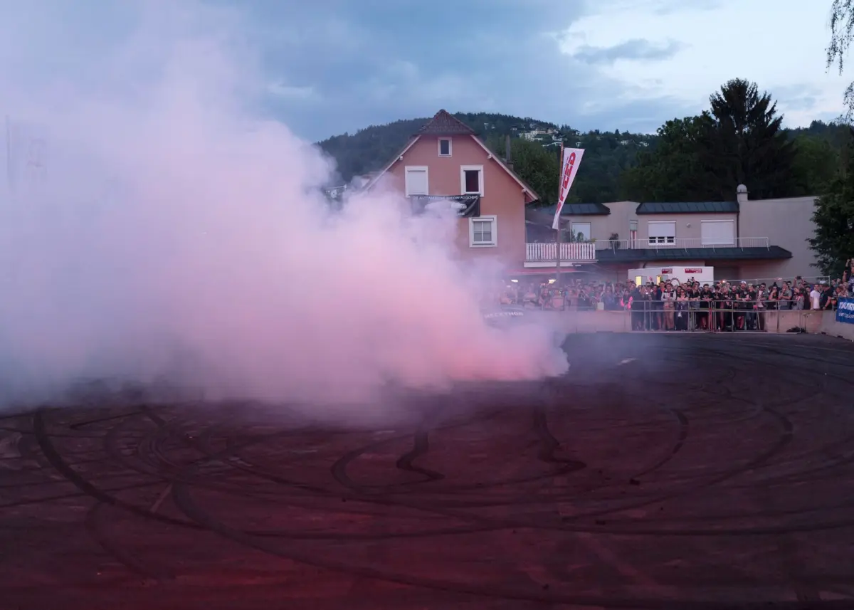 Drifting car emits pink smoke at an outdoor street event; crowd stands behind barriers near a peach-colored house.