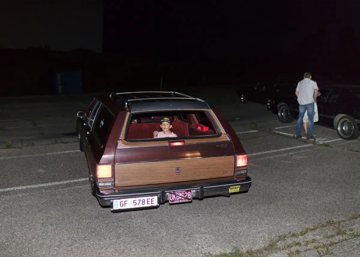 Nighttime parking lot; wood-paneled station wagon in the foreground with a child in the rear seat drinking from a straw.