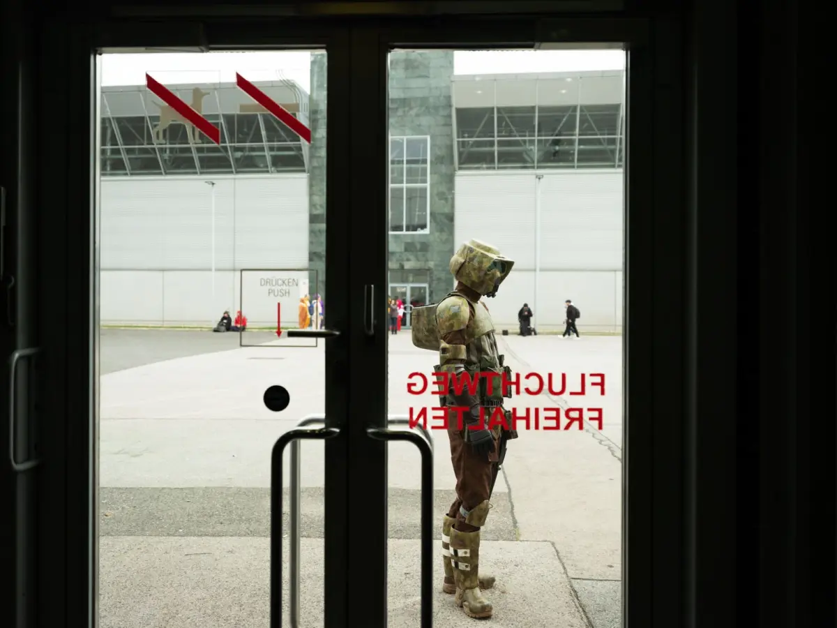 Humanoid robot in camouflage armor stands outside a glass door entrance to a modern building.
