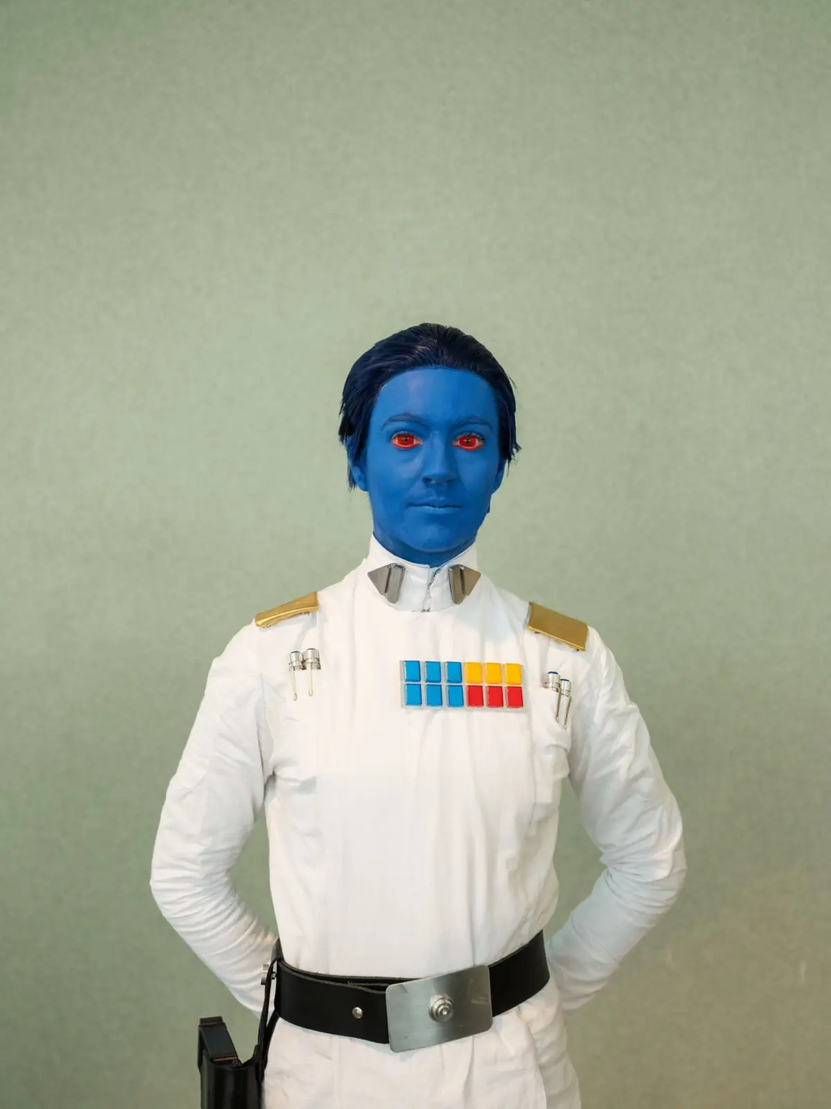 Blue-skinned officer in a white uniform with colorful rank insignia and metallic belt, standing with hands on hips against a pale green wall.