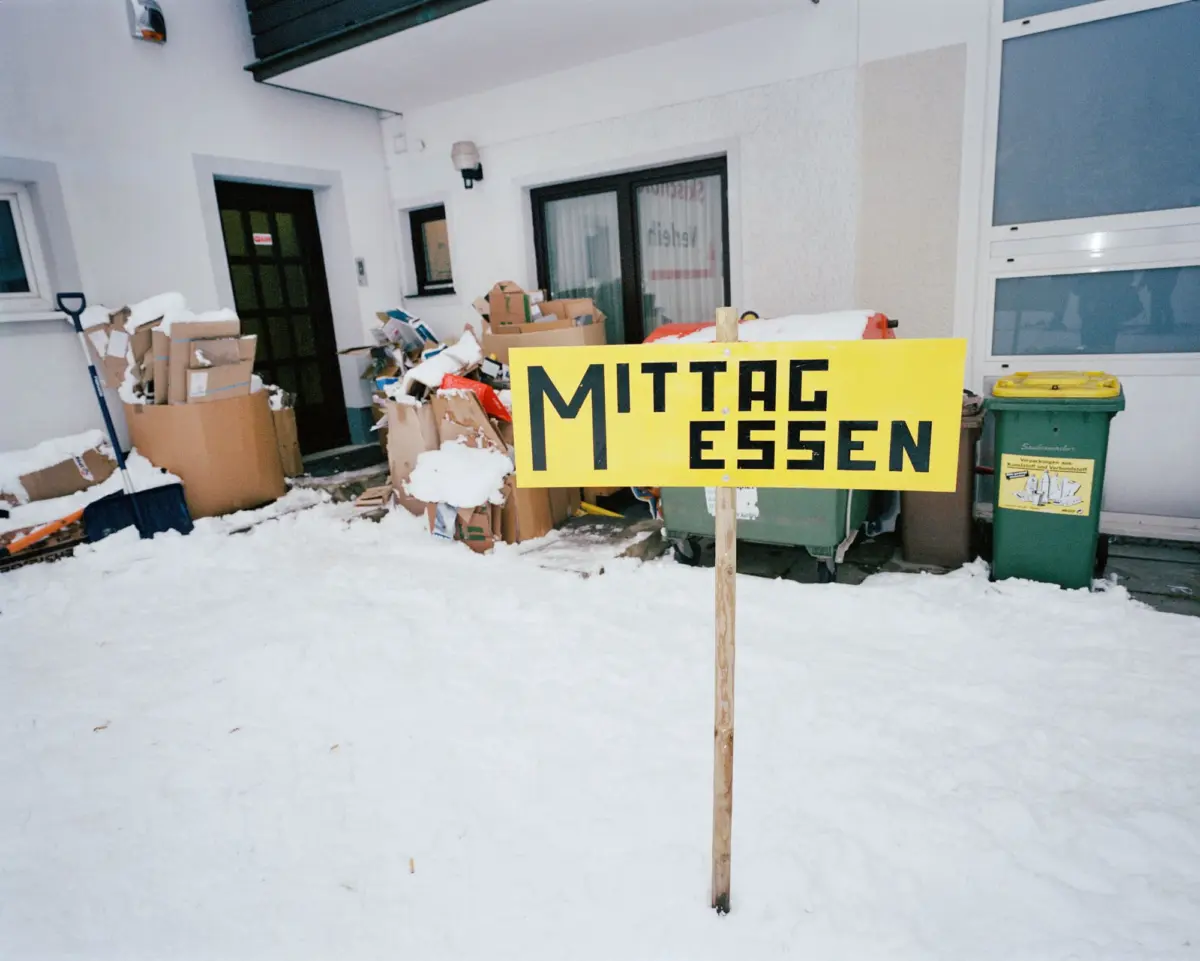 Pile of cardboard boxes and trash in snow by building entrance; bright yellow sign reading MITTAG ESSEN in foreground.