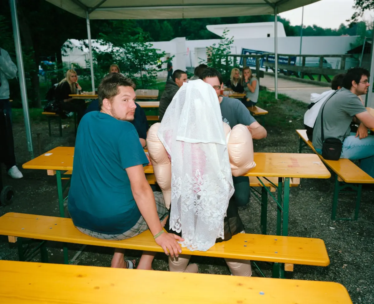 Person wearing a white lace wedding veil stands among adults at an outdoor canopy with yellow picnic tables.