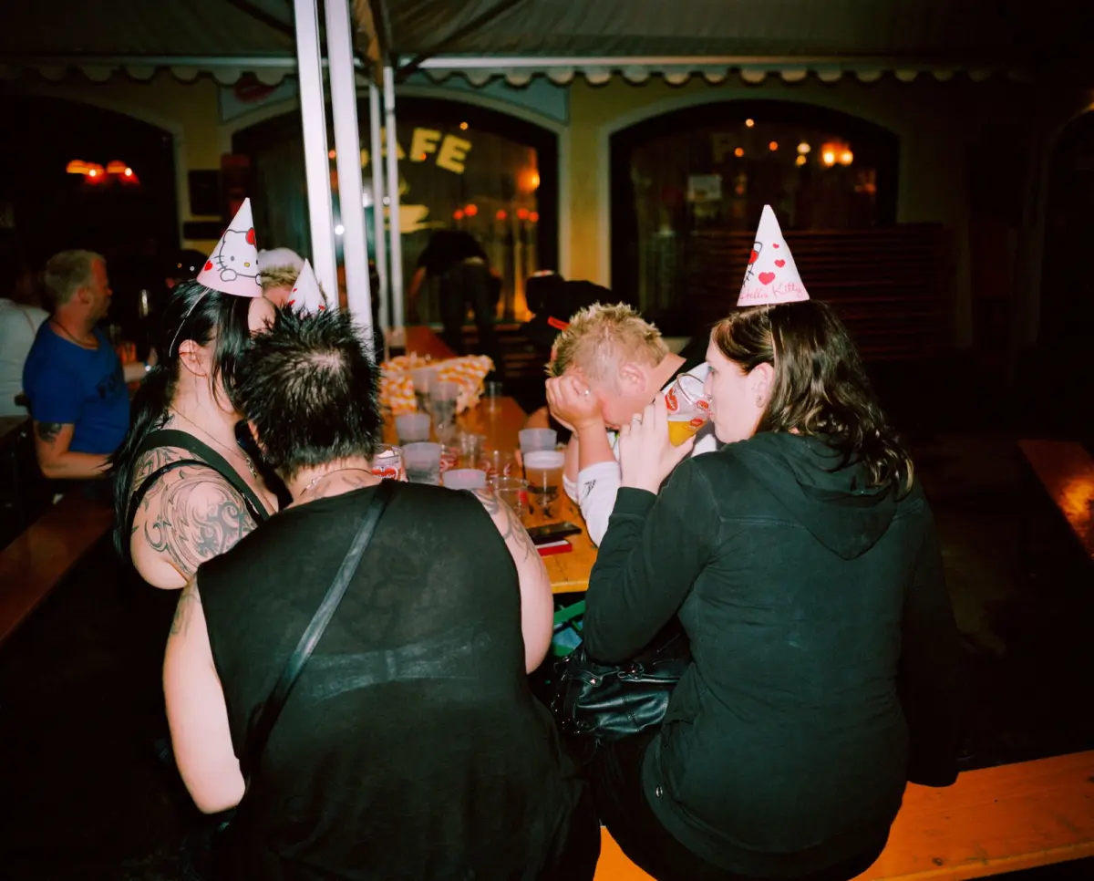 Group of people at a dimly lit bar or cafe, wearing Hello Kitty party hats, drinking and socializing around a long table.