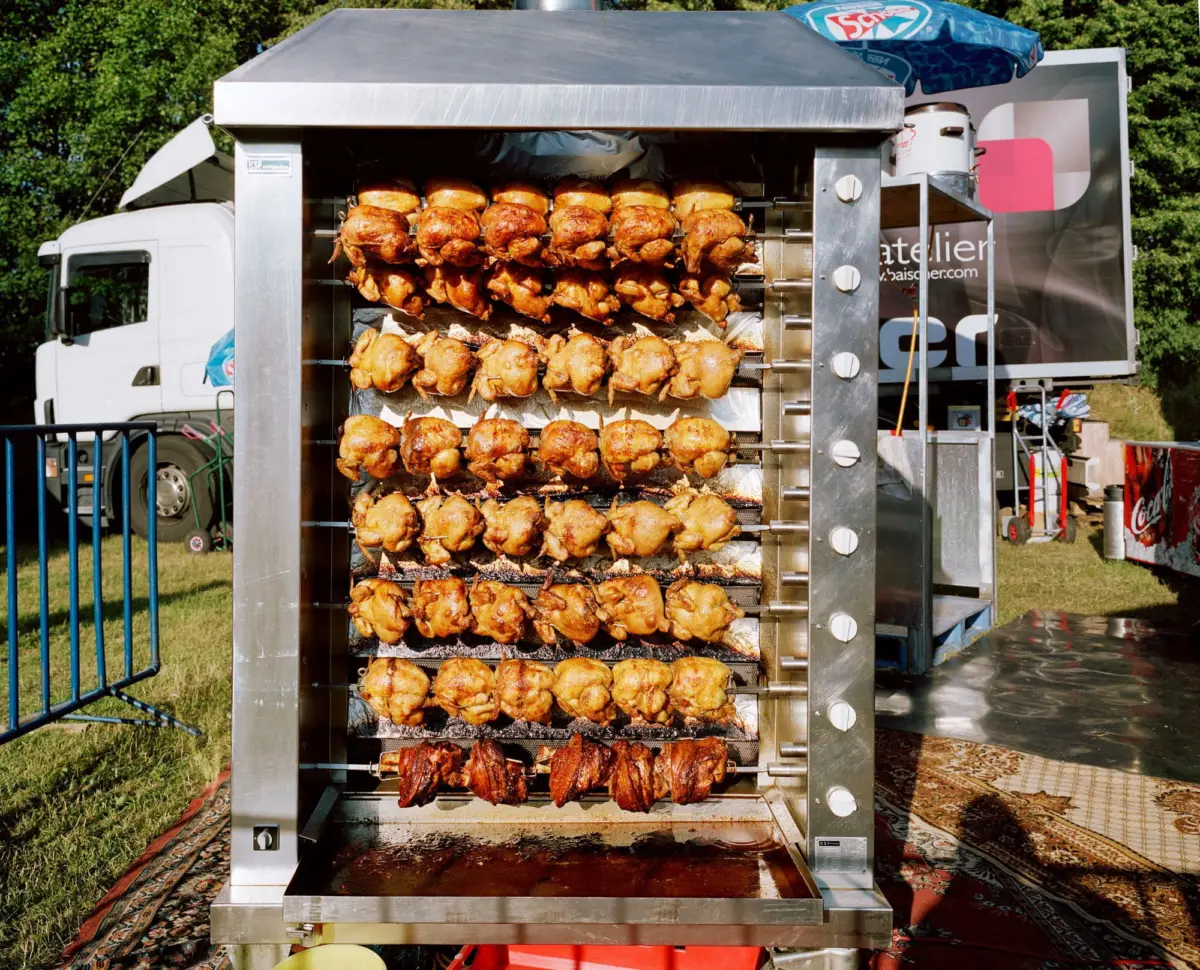 Vertical rotisserie roasting dozens of whole chickens on spits outdoors beside a trailer.