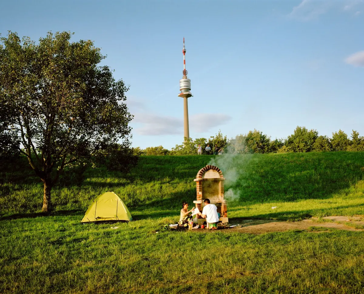 Tall red-and-white cellular tower stands in a grassy park; people cook at a brick grill near a yellow tent.