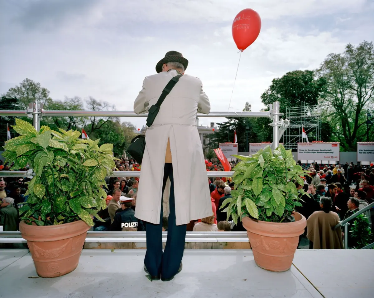 Back view of a person in a beige trench coat and hat standing on a stage with a red balloon, overlooking a crowded outdoor protest.