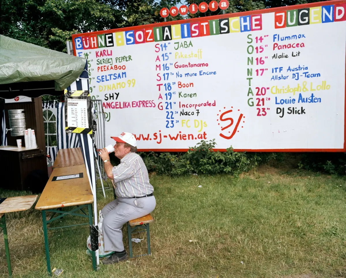 Person in a striped shirt seated at a wooden bench, drinking from a cup, beside a large colorful wall of text at an outdoor festival.