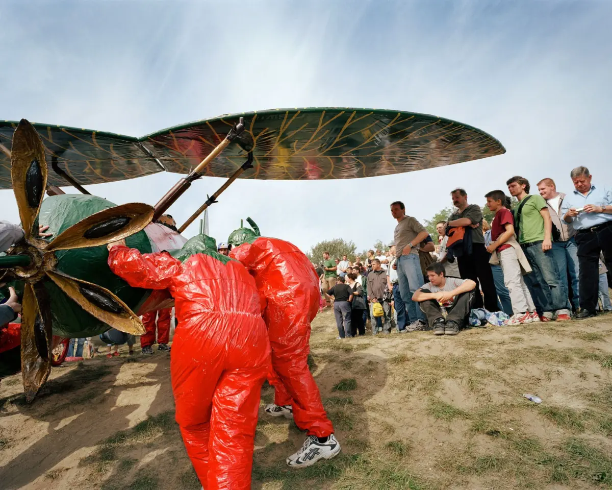 Giant dragonfly-shaped kite with green-veined wings, supported by metal struts, as a crowd watches on a grassy field.