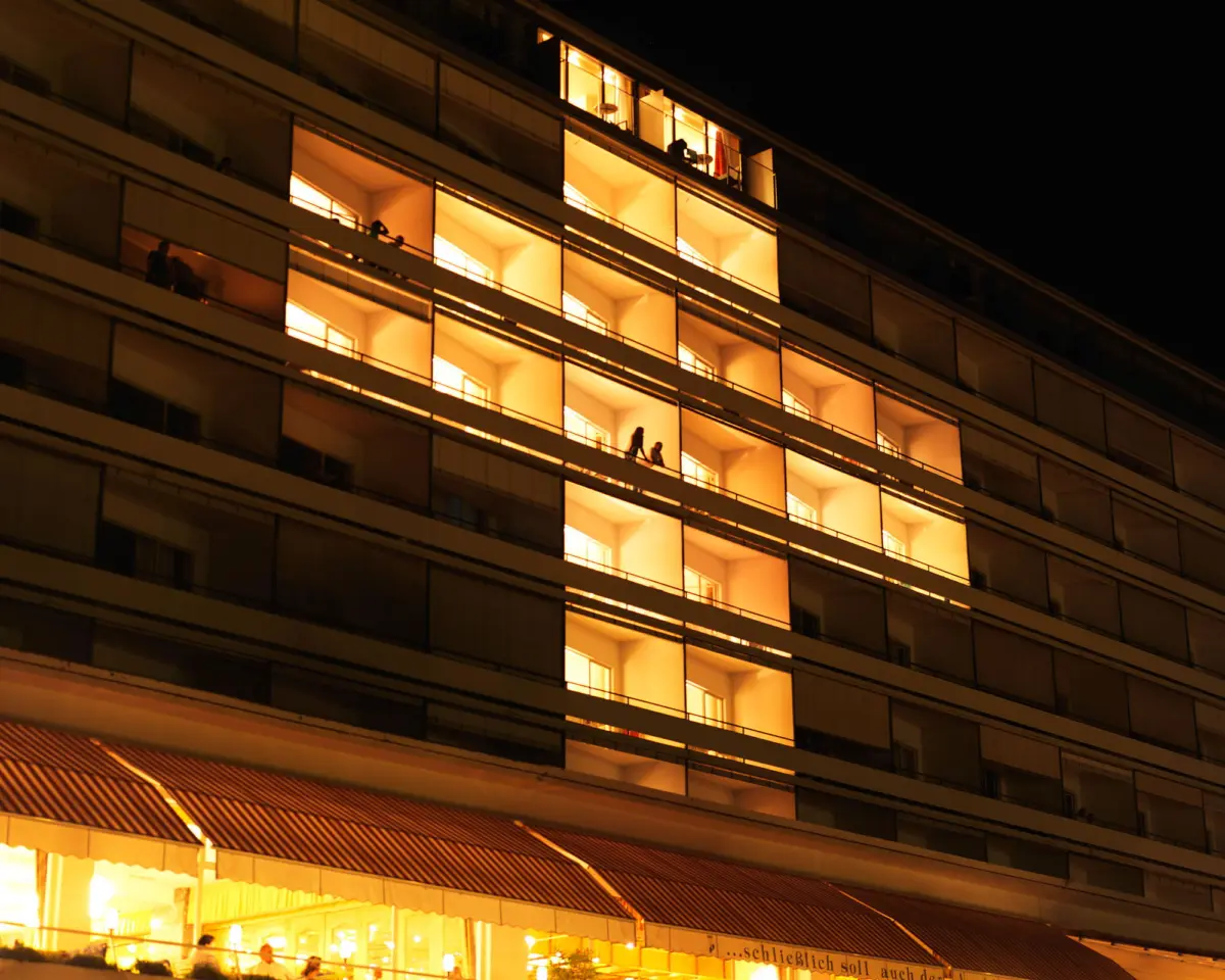 Nighttime view of a multi-story apartment building with warm-lit balconies and silhouetted residents on several levels.