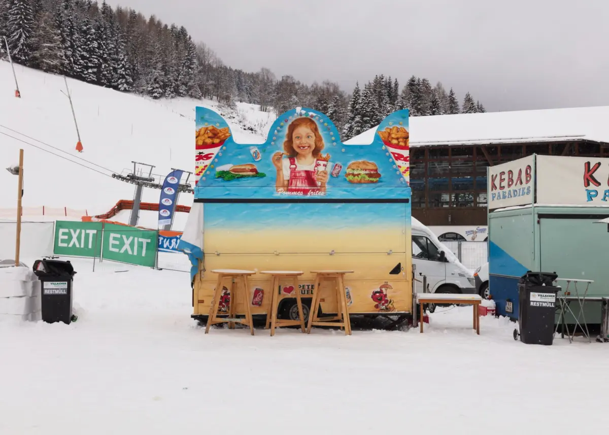 Colorful food trucks parked in a snowy ski resort; blue truck displays a cheerful child image with burgers amid pine trees.