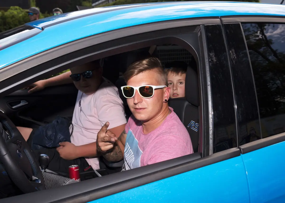 Person in pink shirt with white sunglasses making a hand gesture in a blue SUV; child in back seat looks toward camera.