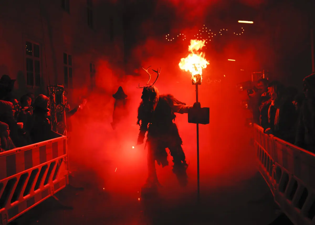 Night street parade; performer in fur cloak and deer antlers carries a flaming torch amid red smoke, crowd behind barriers.
