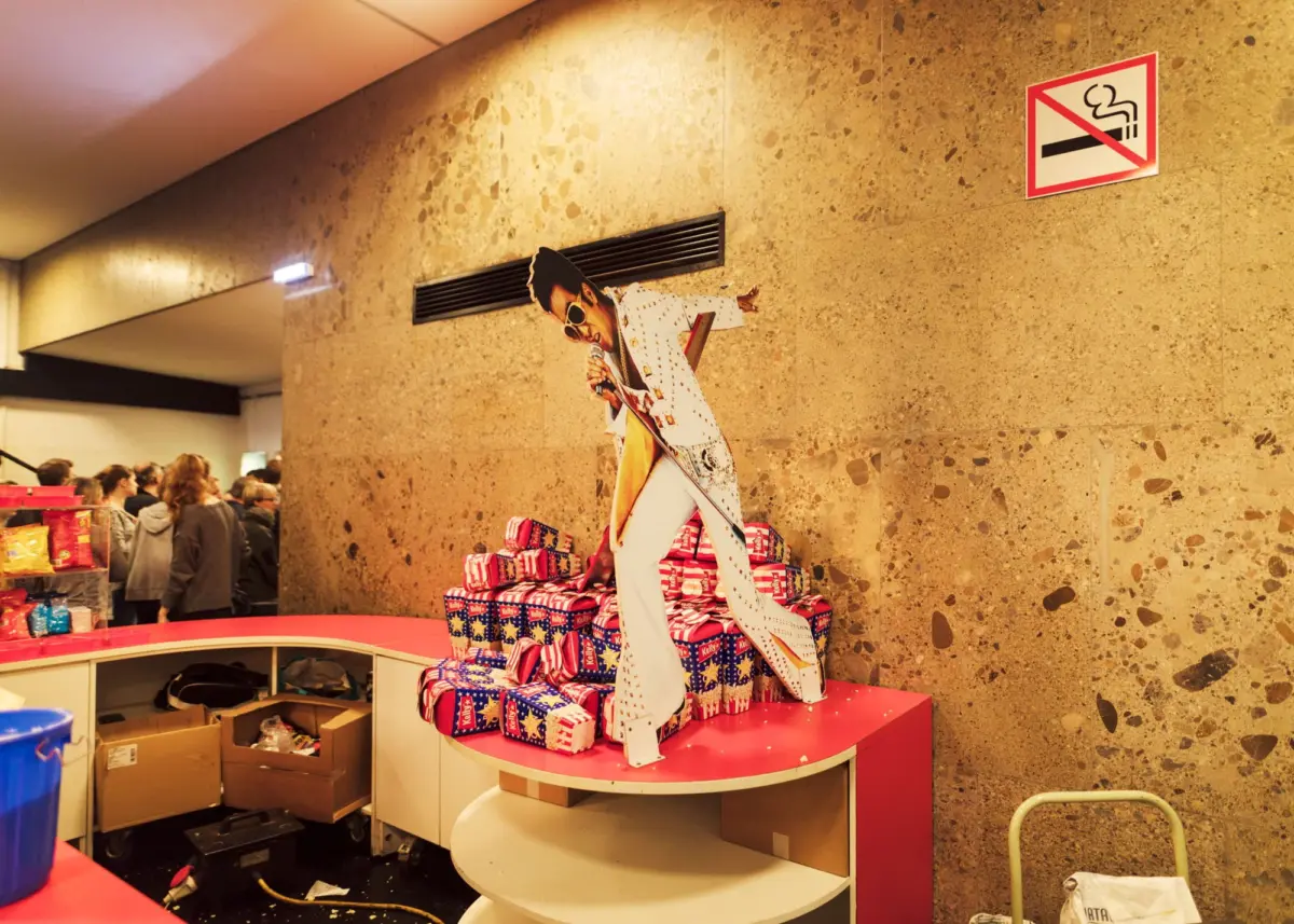 Life-size cardboard cutout of Elvis Presley in a white jumpsuit, leaning by a wall; no-smoking sign and gift-wrapped presents nearby.