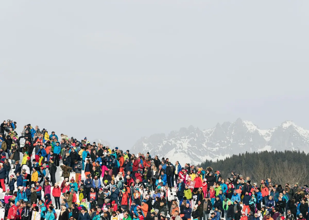 Large crowd of people in colorful winter jackets gathered on a snow-covered mountainside, with snow-capped peaks in the distance.