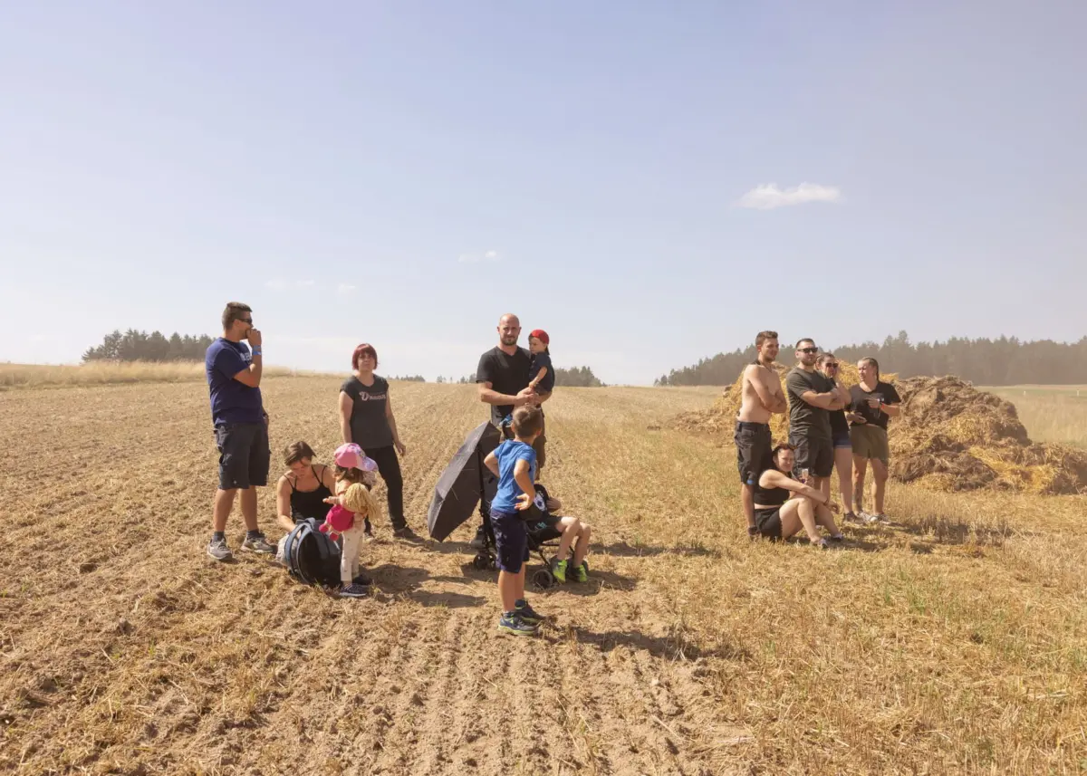Group of adults and children in a sunlit, dry field with haystacks; a man holds a toddler while others watch.