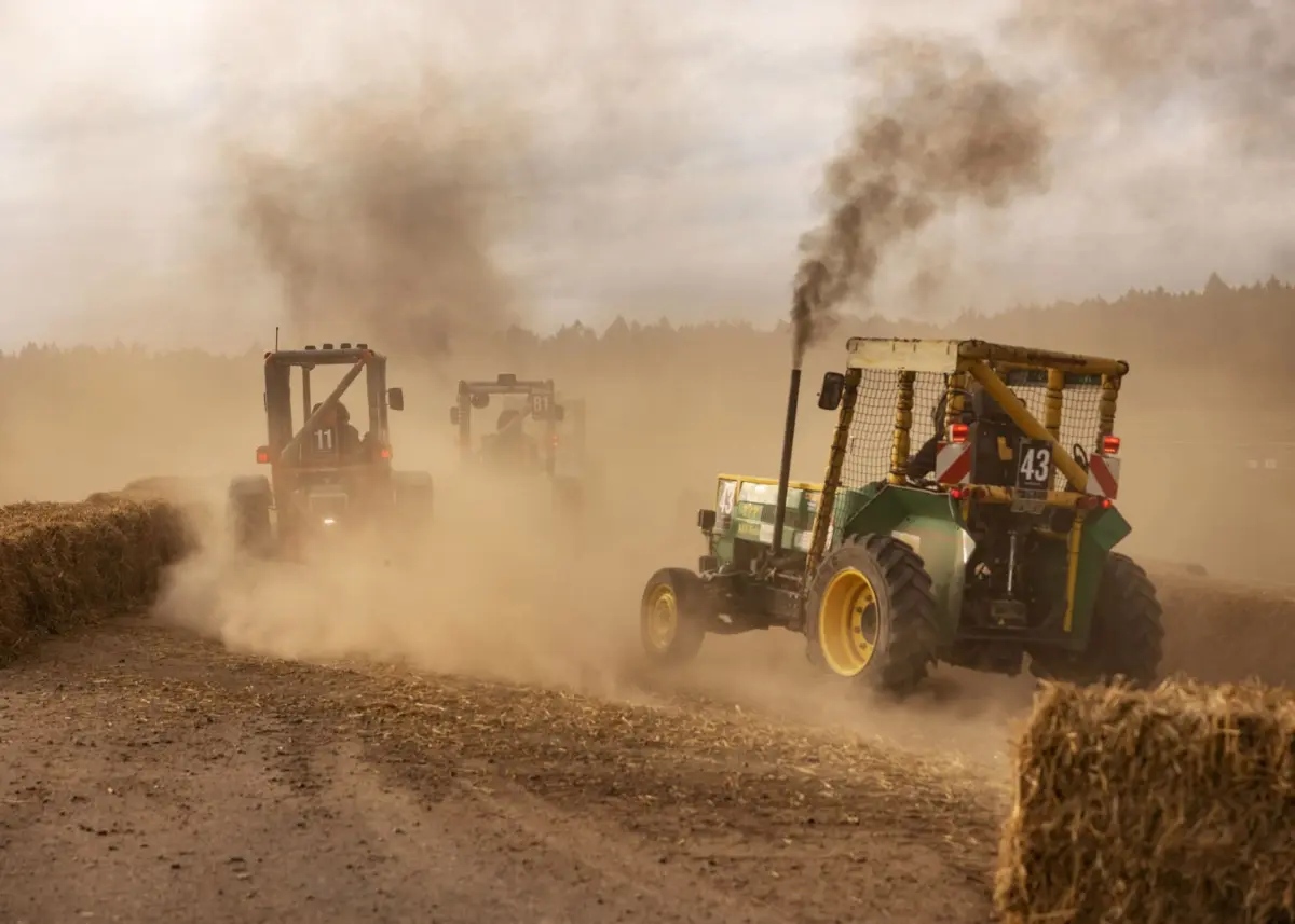Dusty field with green tractors hauling bale trailers; dark exhaust plumes rise as they drive past hay bales.