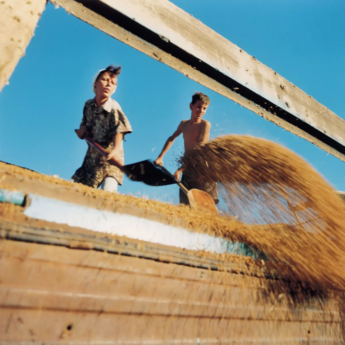 Two young men on a wooden platform, one shirtless and the other in a plaid shirt and white cap, riding past a straw bale under a blue sky.
