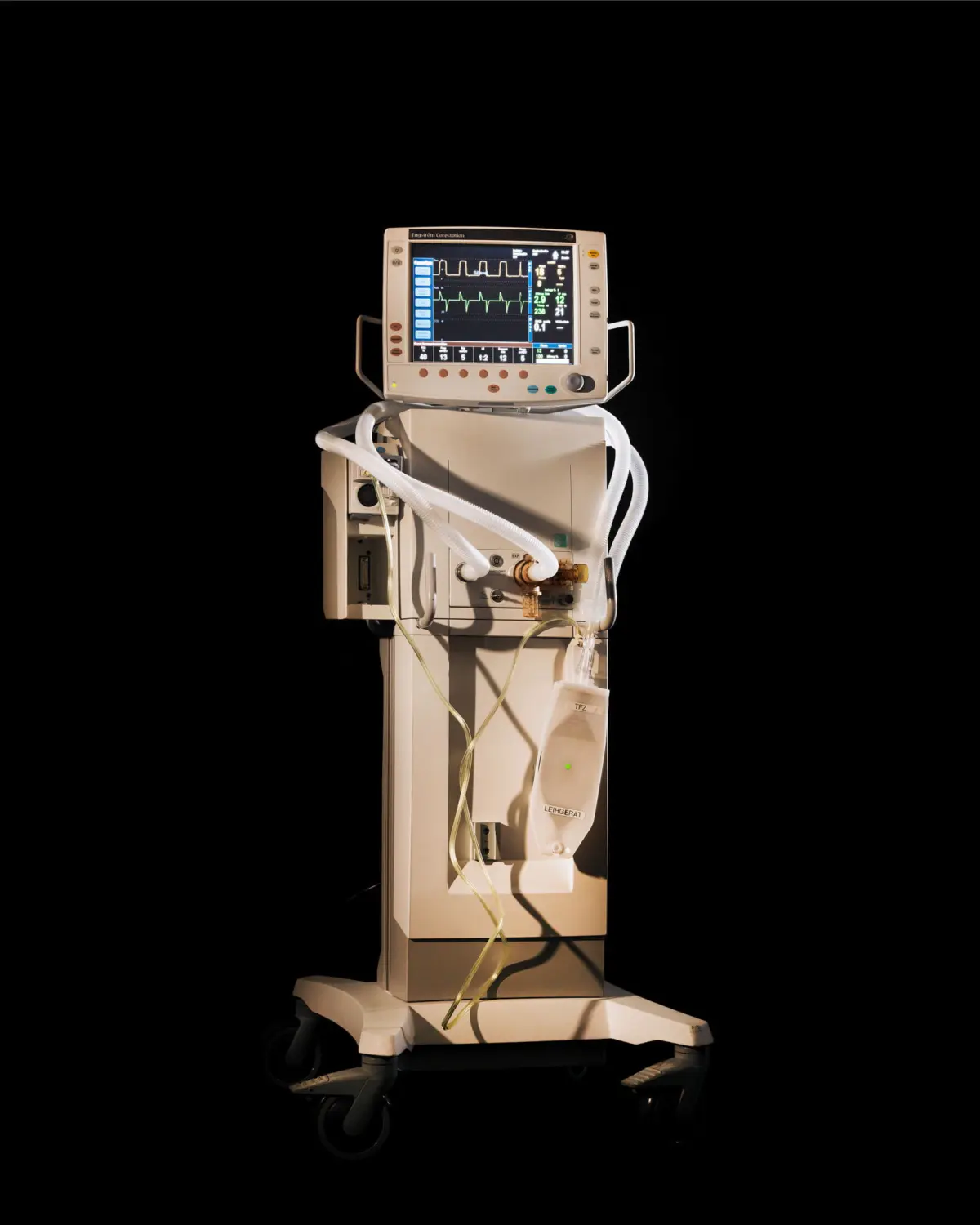 Medical ventilator on a wheeled stand with a monitor displaying vital-sign waveforms against a dark background.