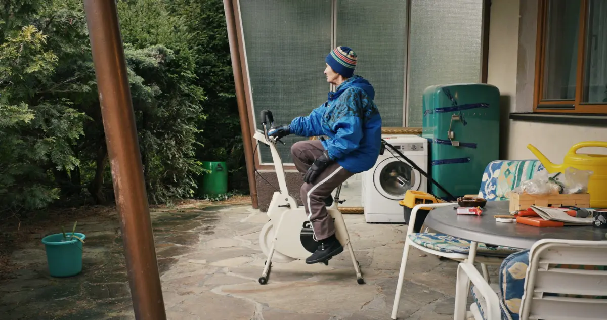 Person on a stationary bike pedaling on a patio beside a washing machine and turquoise refrigerator, with greenery in the background.