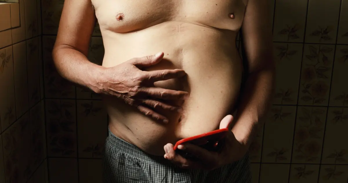 Bare upper body of an adult person, right hand gripping a red object near the abdomen, in a tiled floral bathroom.