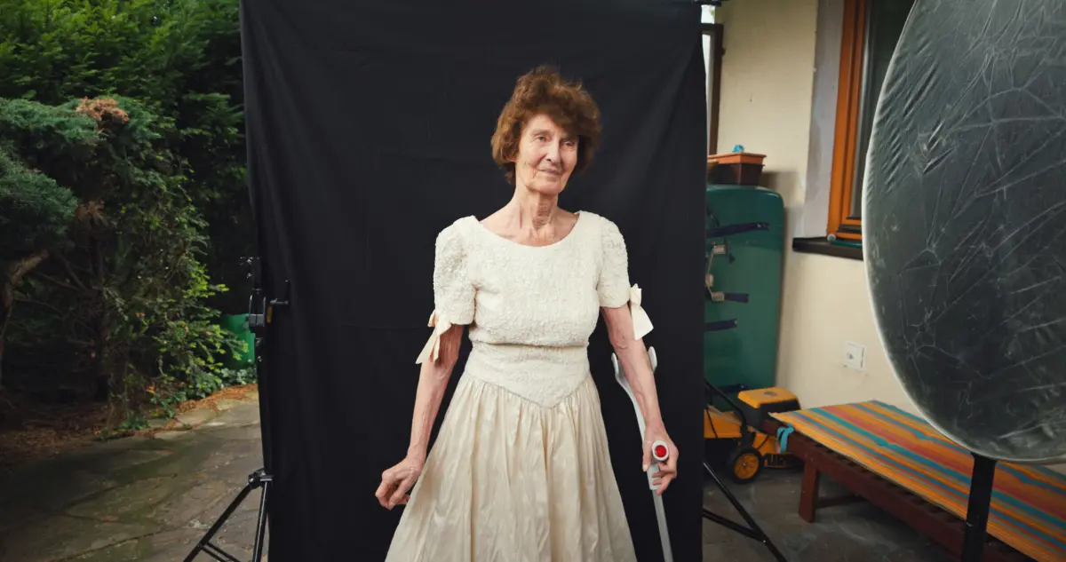 Older woman in ivory lace dress posing for a photo shoot outdoors in a garden with a black backdrop.