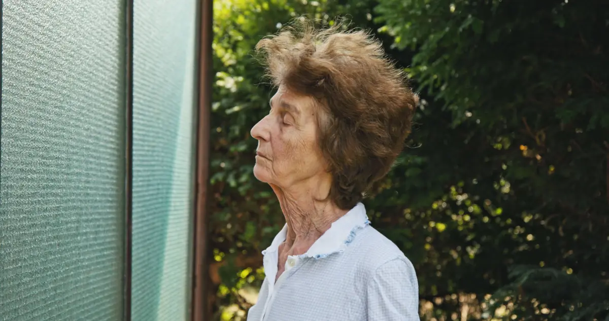 Older woman in a light blue checkered blouse, profile view, standing outdoors beside a textured glass panel with greenery in the background.