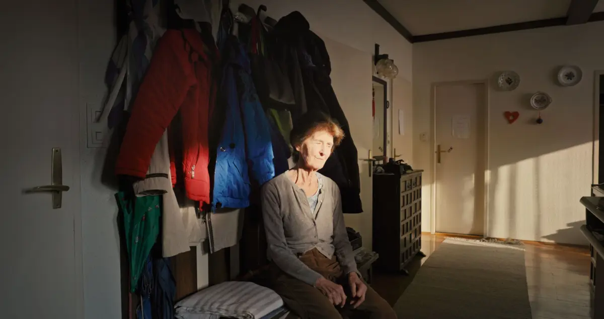 Older person sitting on a bench in a coat-filled entryway, eyes closed, sunlight on the right wall.