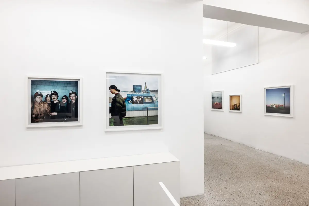 Art gallery with white walls; framed photographs on display, including a group of miners in hard hats and a person beside a blue vehicle by water.