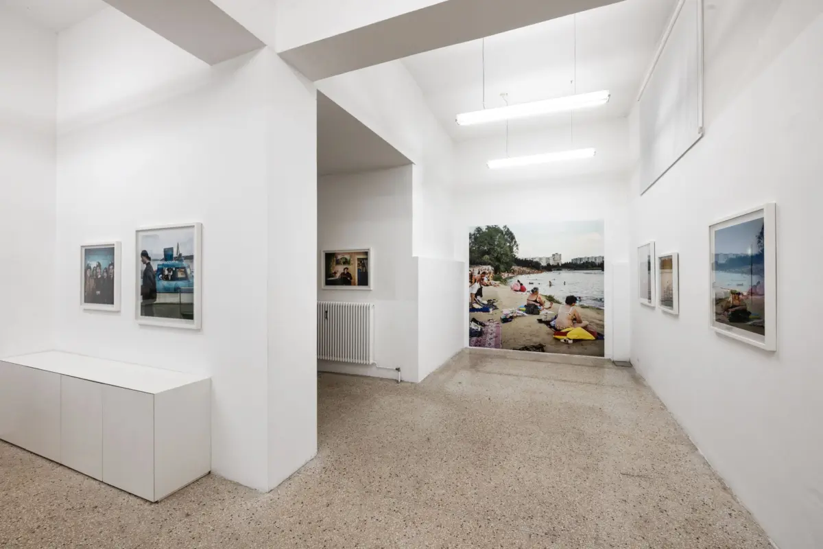 Art gallery interior with white walls and framed photographs, including a large beach scene on the back wall under bright ceiling lights.