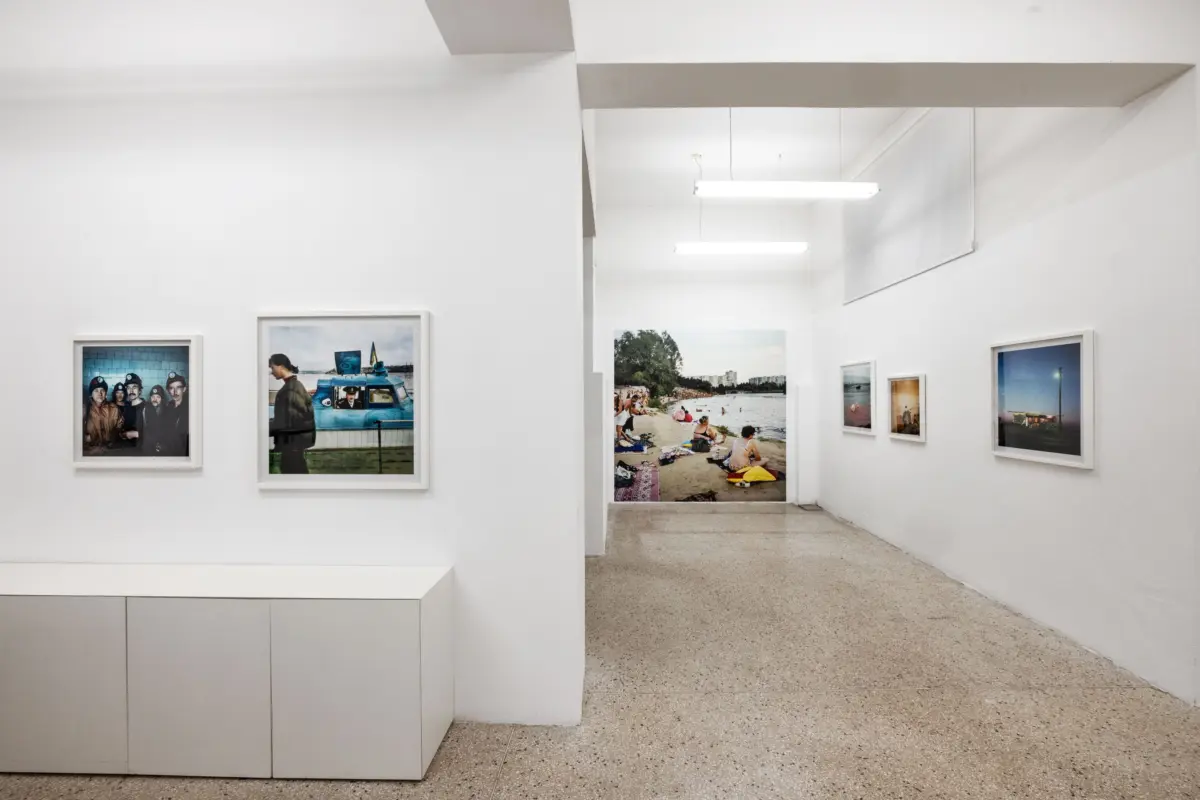 Interior of a white art gallery with framed photographs on display; distant beach scene visible under bright fluorescent ceiling lights.
