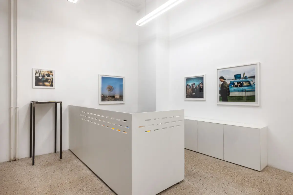 White-walled art gallery with four framed photographs on the walls, a perforated white divider, and a metal display stand.