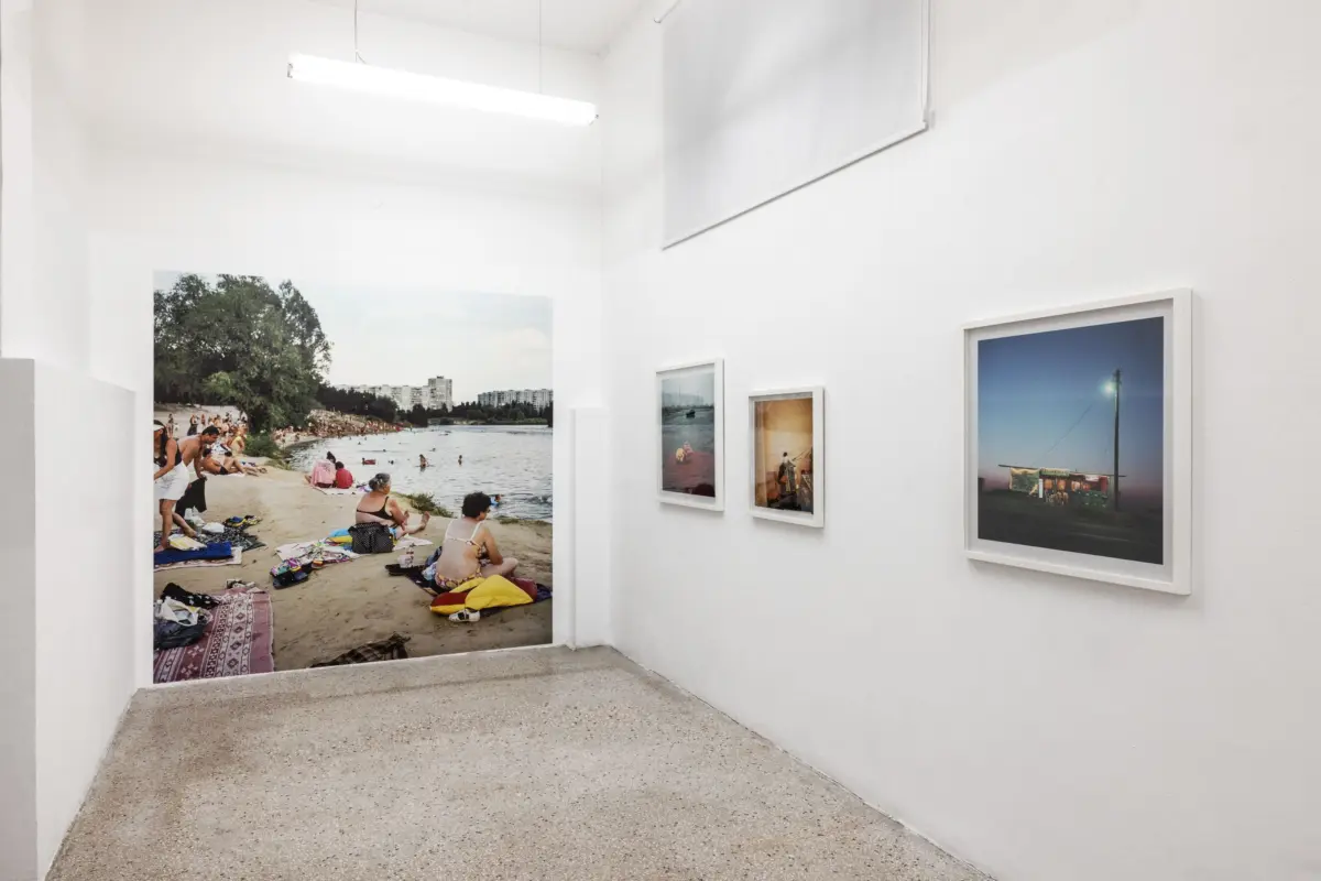Large beach photo of people sunbathing and swimming on a sandy shore with trees and a city skyline, displayed in a white gallery.